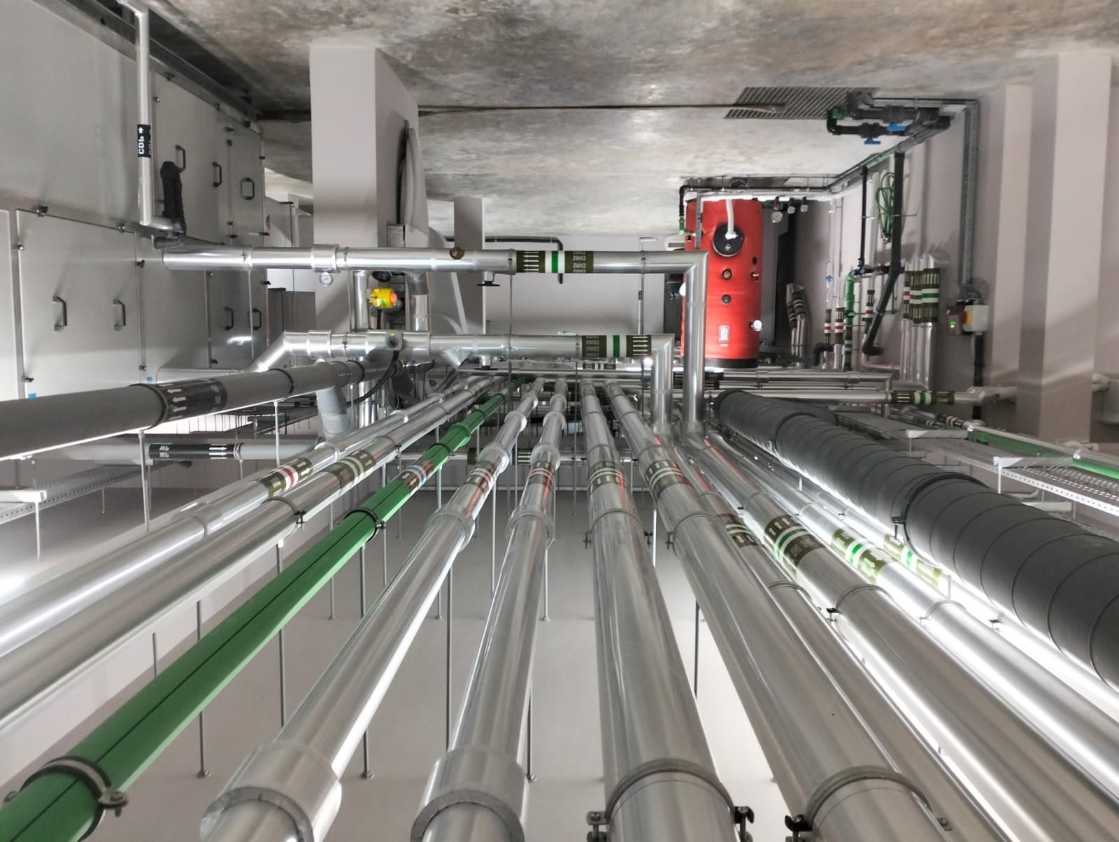 Dense insulated piping runs and air handling units in a building services corridor with a red calorifier vessel
